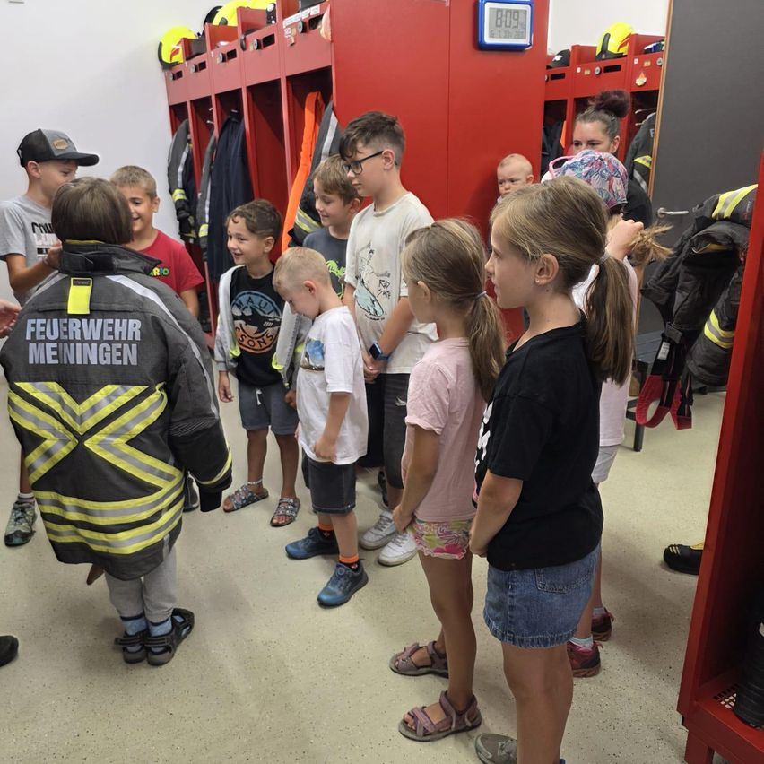 Eine Gruppe von Kindern, einige in Feuerwehruniformen, stehen in einem Raum mit roten Spinden und einer digitalen Uhr. Sie schauen einen Feuerwehrmann an.