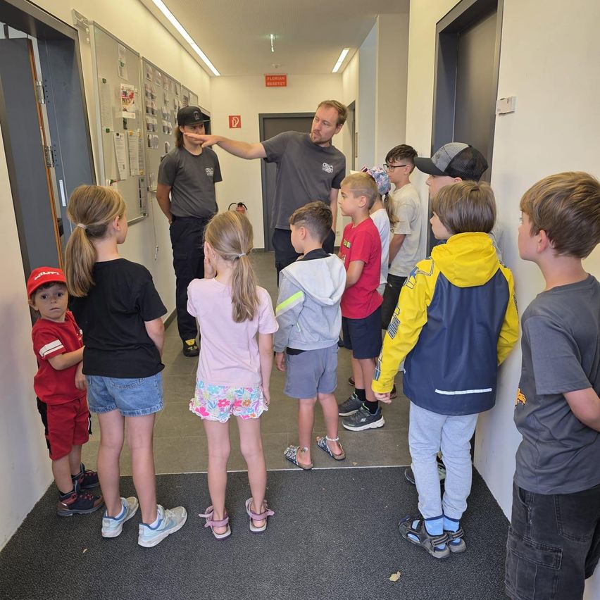 Eine Gruppe von Kindern und zwei Erwachsenen steht in einem Flur und hört einem Mann zu, der auf eine Wand zeigt.