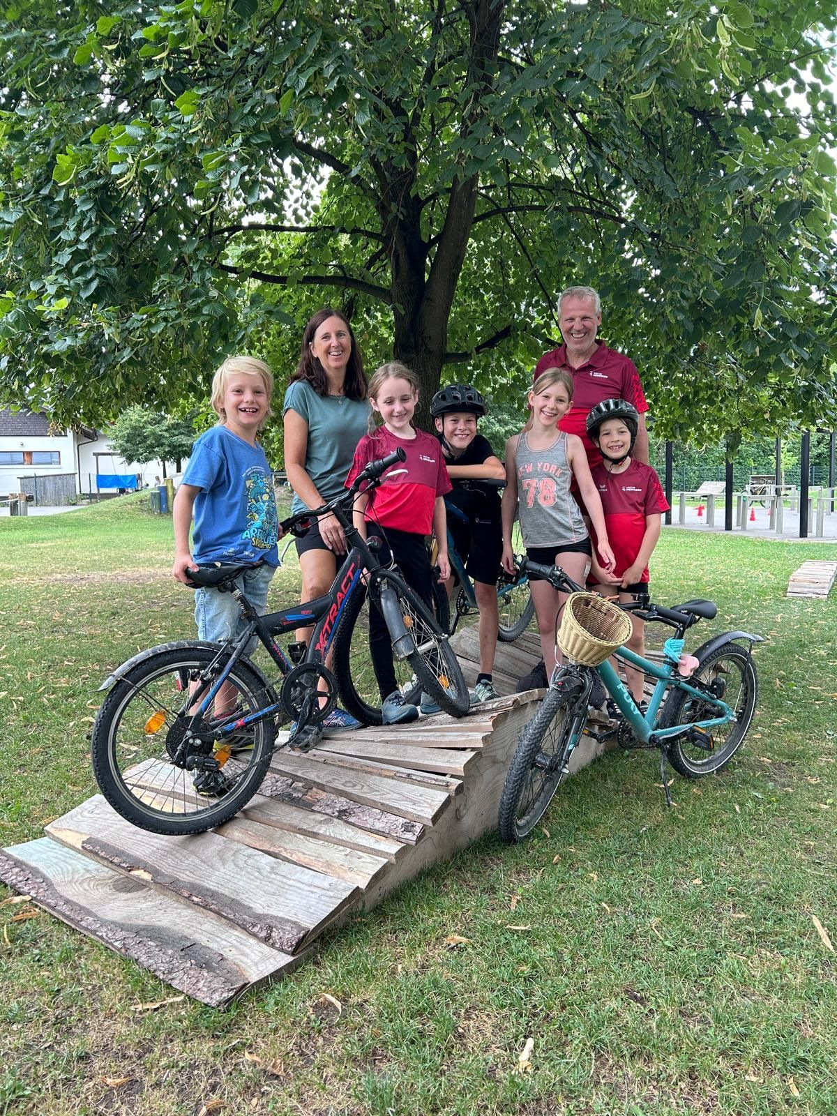 Eine Familie mit sieben Personen, darunter Kinder, steht unter einem Baum mit ihren Fahrrädern auf einer Holzrampe. Sie alle lächeln und scheinen für ein Foto zu posieren.