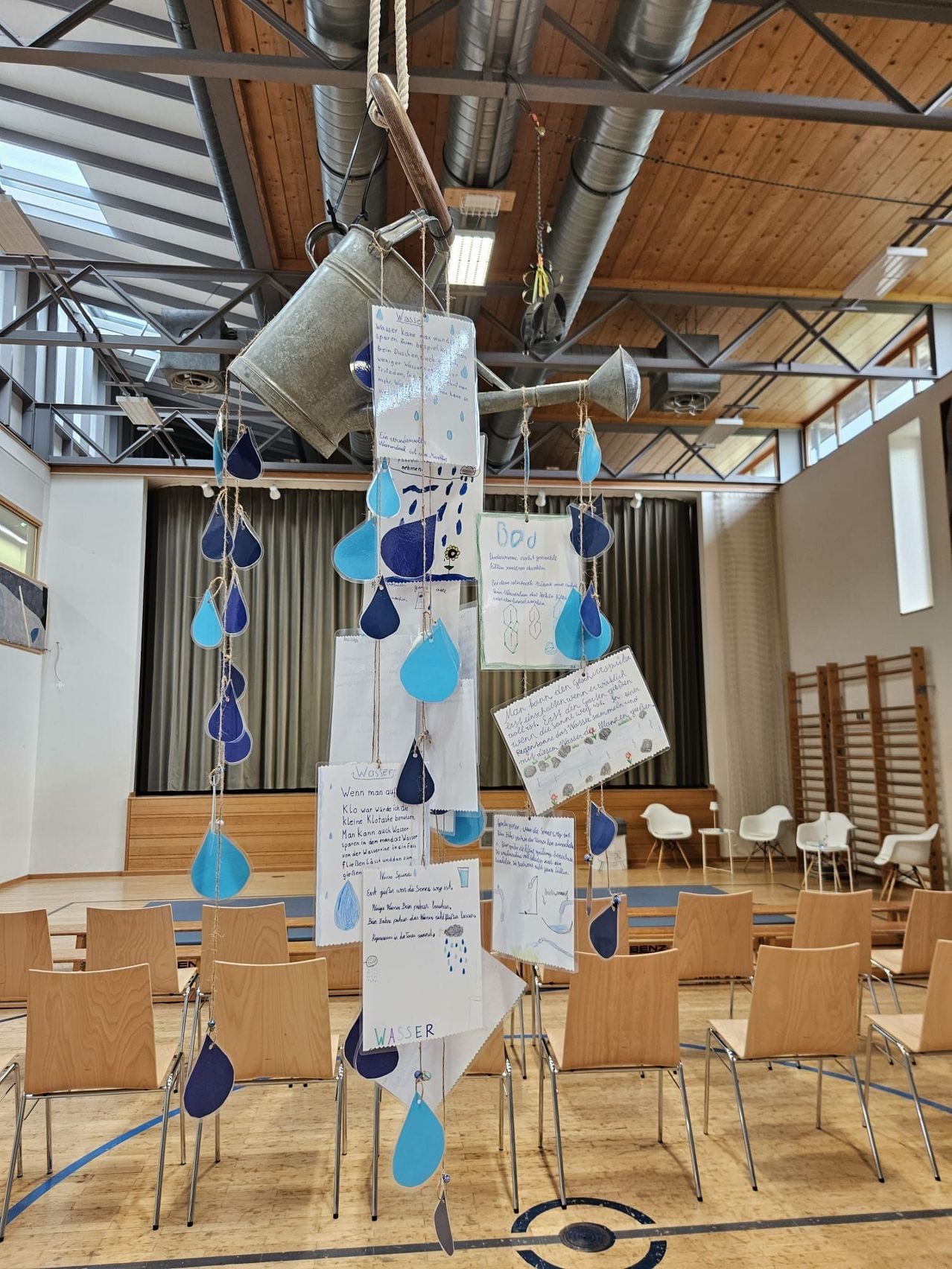 Ein Regentropfen-förmiges Mobile hängt in einem großen Raum mit Holzstühlen. Das Mobile hat blaue Tröpfchen mit Text. Ein Metalleimer hängt von der Decke. Ein weißer Vorhang befindet sich hinter den Stühlen.