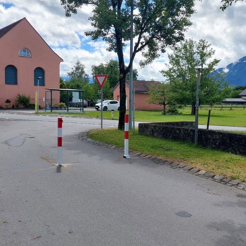 Eine Straße mit rot-weißen Pfosten, einem rot-weißen dreieckigen Schild und einem rosa Gebäude mit Bushaltestelle und geparkten Autos im Hintergrund.