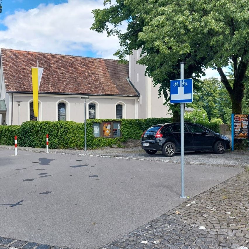 Ein schwarzes Auto ist vor einem Gebäude mit einem braunen Dach und mehreren Fenstern geparkt. Im Vordergrund befindet sich ein Fahrradschild.
