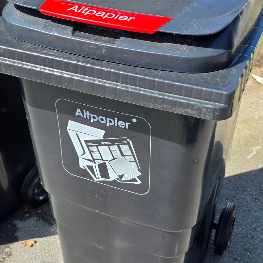 Ein schwarzer Recyclingbehälter mit einem roten Deckel, der mit 'Altpapier' beschriftet ist. Der Körper des Behälters hat einen weißen Aufkleber mit demselben Text und einer Illustration gefalteter Papiere.