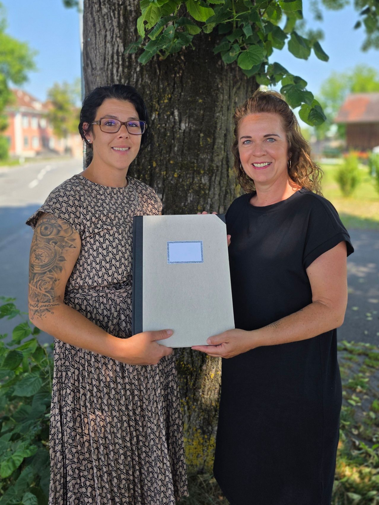 Two women stand outside, smiling and holding a book, with one having a tattoo and both standing by a tree.