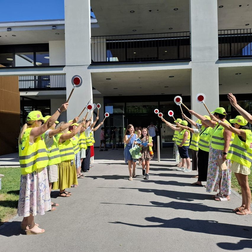 A group of people in yellow vests and hats are holding red and white flags in front of a building. A girl and a woman are walking in the middle.