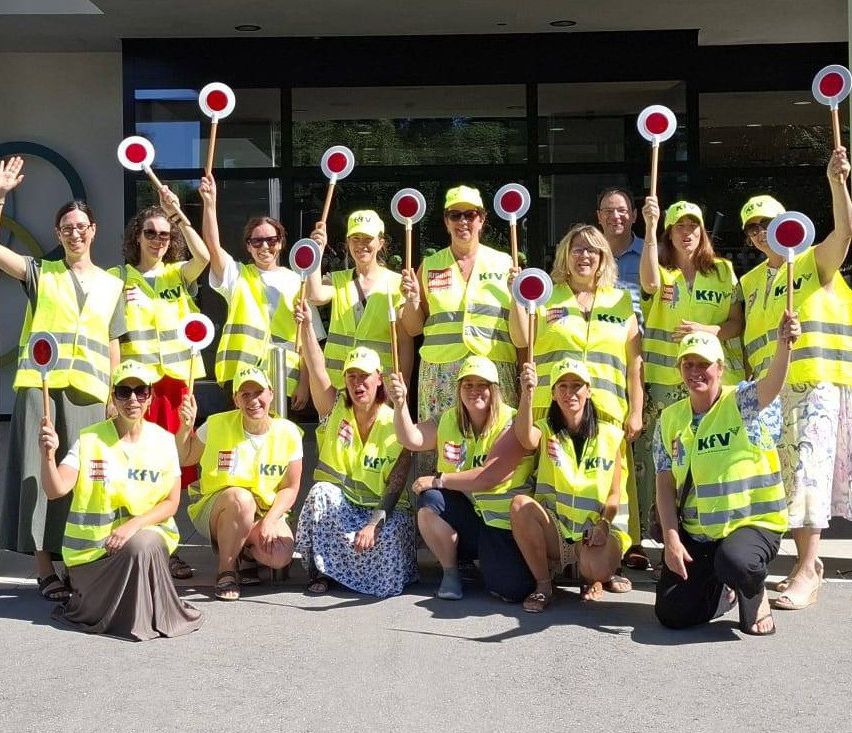A group of volunteers in yellow vests and hats, holding red and white circular signs, pose for a photo in front of a building.