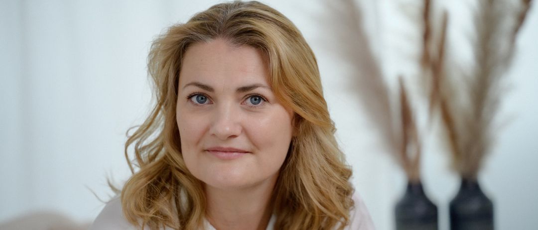 Eine Frau mit blonden Haaren lächelt in die Kamera. Sie hat blaue Augen und trägt ein weißes Hemd. Der Hintergrund ist hell.