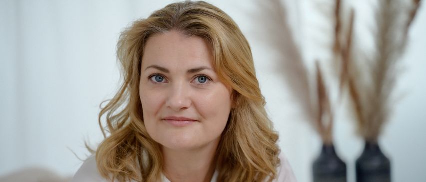 Eine Frau mit blonden Haaren lächelt in die Kamera. Sie hat blaue Augen und trägt ein weißes Hemd. Der Hintergrund ist hell.