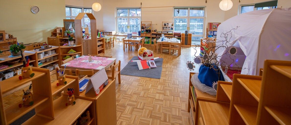 Ein Kindergartenraum mit Holzregalen, Tischen, Stühlen und einer Holzdecke. Helles Licht fällt durch die Fenster. Ein Teppich mit einem Teddybären und Büchern liegt auf dem Boden.