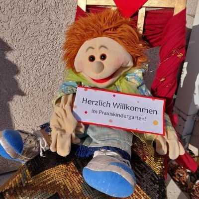 Eine Puppe sitzt auf einem Stuhl mit einem roten Herz darüber und hält ein Schild mit der Aufschrift 'Herzlich Willkommen im Praxiskindergarten!' Die Puppe trägt blaue Turnschuhe und ein grünes Halstuch.