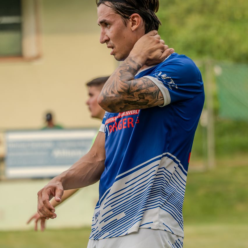 Ein Mann in einem blauen Fußballtrikot mit einer Tätowierung am Arm steht auf einem Rasenfeld und reibt sich den Nacken.
