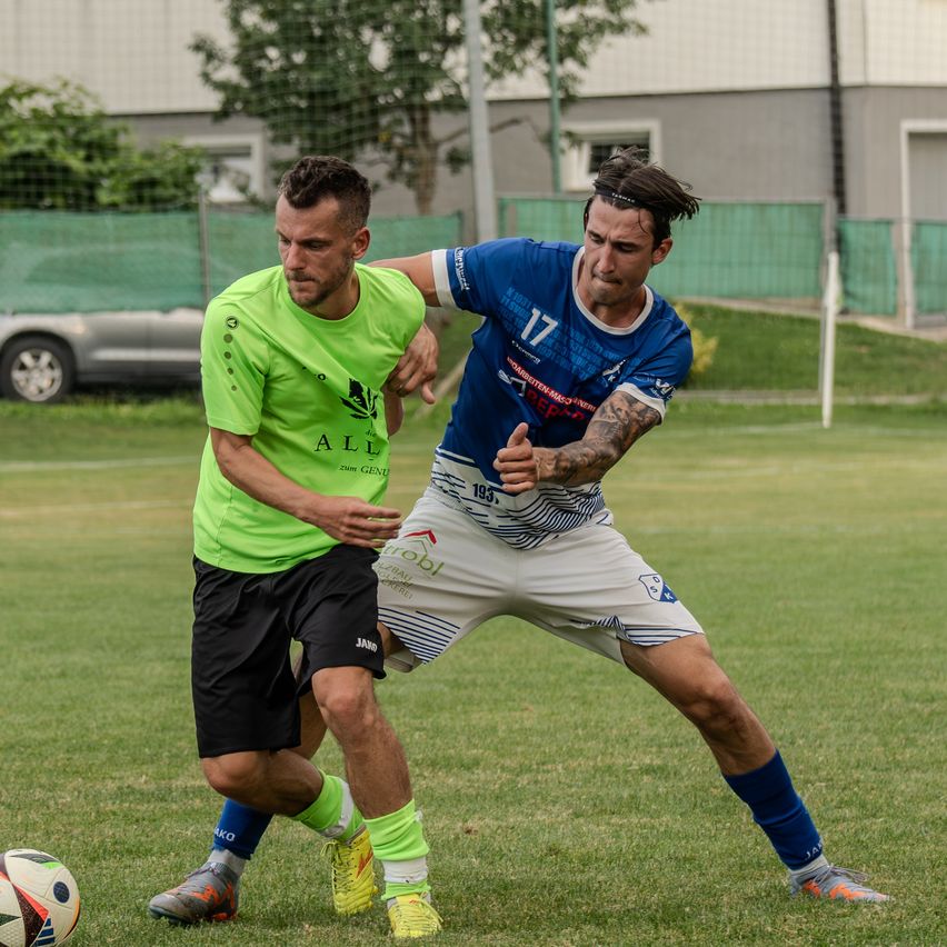 Zwei Fußballspieler konkurrieren auf einem Rasenfeld. Einer trägt ein grünes Trikot, der andere ein blaues Trikot mit der Nummer 17. Sie versuchen beide, die Kontrolle über den Ball zu erlangen.