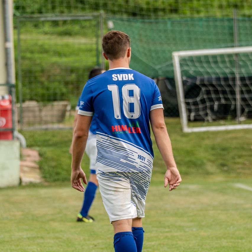 Ein Fußballspieler mit der Nummer 18 auf einem blauen Trikot läuft auf einem Rasenfeld. Dahinter ist eine verschwommene Ansicht eines anderen Spielers und eines Tors.