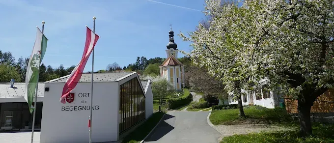 Ein weißes Gebäude mit Flaggen steht in einer grünen Landschaft. Ein Weg führt zu einem Kirchturm. Der Himmel ist blau und klar.