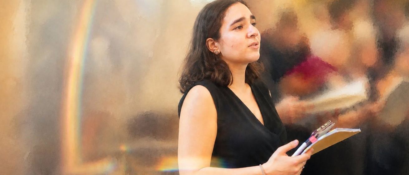 Eine Frau mit lockigem Haar, die ein schwarzes ärmelloses Oberteil trägt, eine Mappe in der Hand hält und mit einem Regenbogen im Hintergrund in die Kamera schaut.