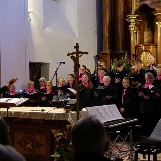 Ein Chor singt in einer Kirche, mit Frauen in rosa Schals, einem Holzkreuz und einem Klavier auf der Bühne. Das Publikum sitzt unten und schaut zu.