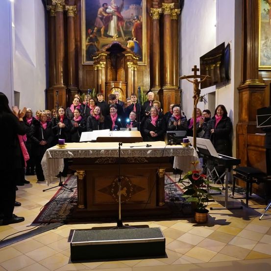 Ein Chor in pink steht vor einem Altar in einer Kirche. Dahinter steht ein Kreuz auf einem Ständer, und ein Klavier und ein Mikrofon sind rechts aufgestellt.