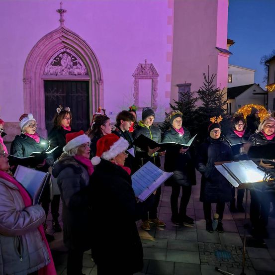 Ein Chor in Weihnachtsmützen singt vor einer nachts in Pink beleuchteten Kirche.