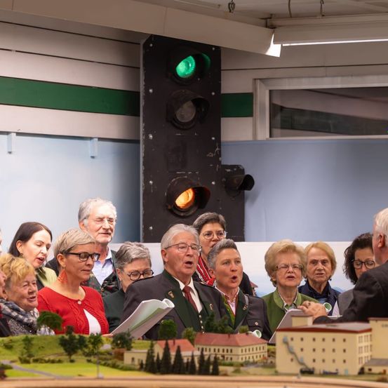 Eine Gruppe älterer Erwachsener, einige mit Brille, singen vor einer Modelleisenbahnaufstellung, während eine Ampel mit grüner Ampel im Hintergrund steht.
