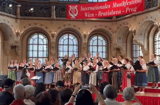 Ein Chor tritt auf der Bühne bei einer Veranstaltung auf, mit einem Banner oben, das Internationales Musikfestival Wien-Bratislava-Prag liest. Zuschauer sitzen im Vordergrund.