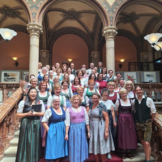 Eine Gruppe von Menschen, gekleidet in traditioneller deutscher Kleidung, posiert für ein Foto auf einer großen Treppe. Die Architektur ist üppig, mit Säulen und dekorativen Elementen.