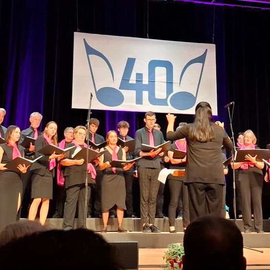 Ein Chor tritt auf der Bühne auf und hält Notenblätter in den Händen. Sie tragen einheitliche Kleidung und über ihnen hängt ein Schild mit Musiknoten und '40'.
