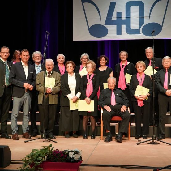 Eine Gruppe von Menschen in formeller Kleidung steht auf einer Bühne mit Mikrofonen und Blumen. Sie halten Urkunden und scheinen ein Ereignis zu feiern. Ein Banner hinter ihnen trägt die Aufschrift '40'.