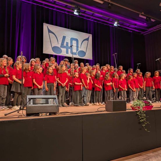 Ein Chor in roten Kleidungsstücken steht auf der Bühne, mit einem Banner über ihnen, das '40' lautet. Mikrofone und Lautsprecher sind aufgestellt. Sie werden von lila Bühnenlichtern beleuchtet.