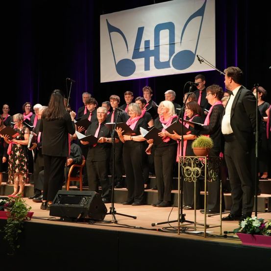 Ein Chor aus Männern und Frauen in schwarzer und rosa Kleidung tritt mit Mikrofonen und Notenständern auf der Bühne auf, über ihnen ein Schild mit der Aufschrift '40'.