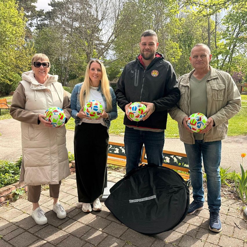 Vier Menschen in Outdoor-Kleidung posieren mit bunten Fußballen. Hinter ihnen steht eine Bank und ein Beutel auf dem Boden. Im Hintergrund sind Bäume und ein klarer Himmel zu sehen.