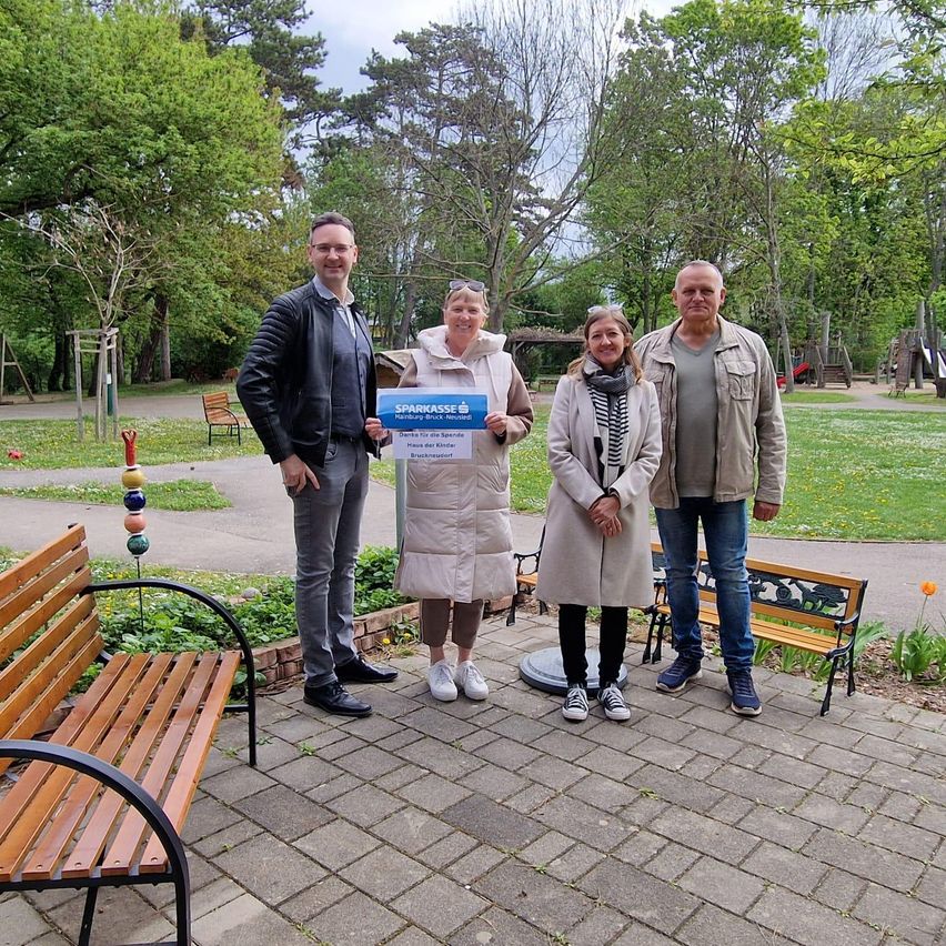 Vier Personen posieren in einem Park. Eine Frau hält ein blaues Banner. Eine Bank und Bäume sind im Hintergrund.