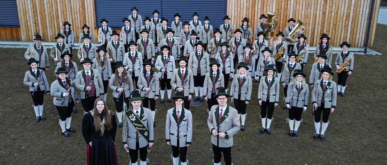 Eine Gruppe von Musikern, in traditioneller Kleidung gekleidet, steht in einer Reihe und hält ihre Instrumente vor einem Holzgebäude.