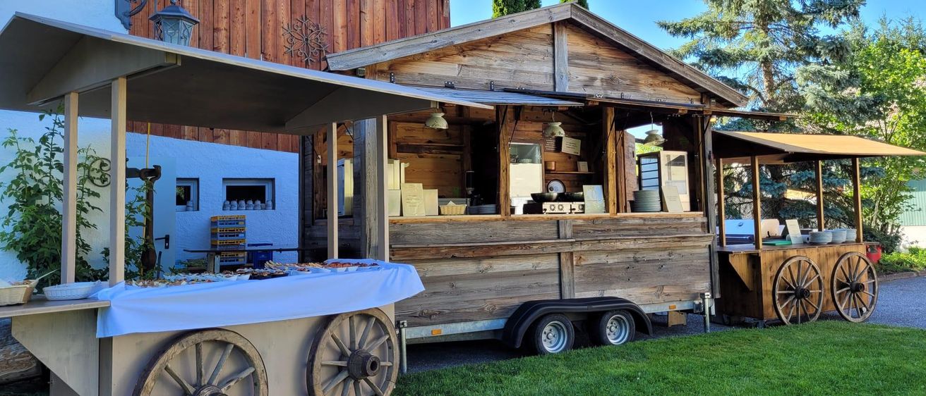 Ein Holzimbisswagen steht vor einem Holzhaus mit grünem Rasen. Vor ihm steht ein Tisch mit einem weißen Tuch.
