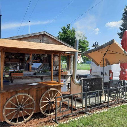 Ein rustikaler Holzimbisswagen mit zwei großen Rädern steht auf einem Erdboden. Ein Mann kocht im Inneren, und Rauch kommt aus dem Schornstein. Ein Regenschirm ist neben dem Wagen aufgestellt.
