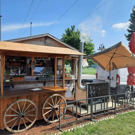 Ein rustikaler Holzimbisswagen mit zwei großen Rädern steht auf einem Erdboden. Ein Mann kocht im Inneren, und Rauch kommt aus dem Schornstein. Ein Regenschirm ist neben dem Wagen aufgestellt.
