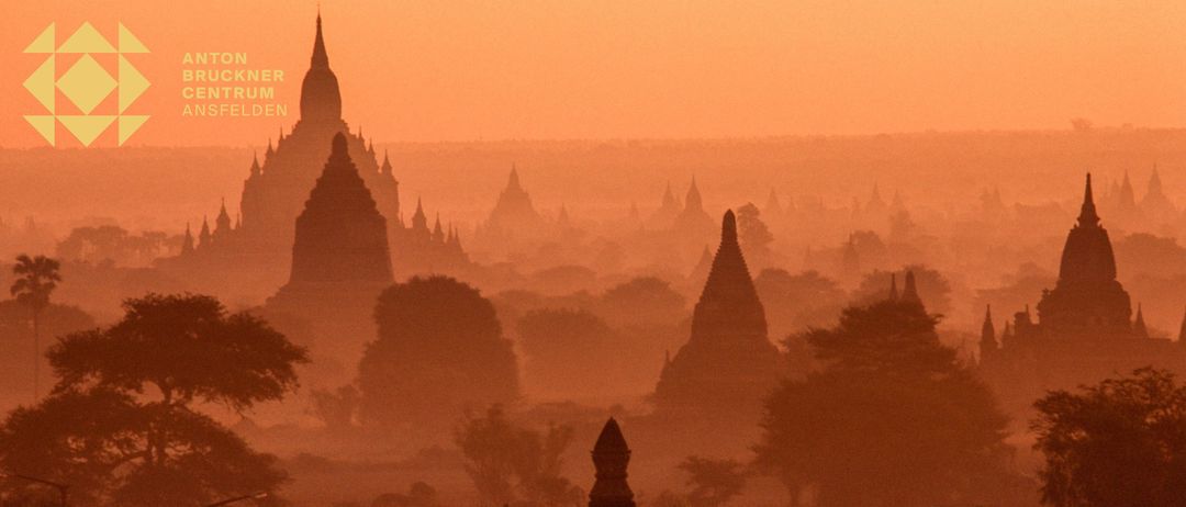 Eine neblige Landschaft mit alten Pagoden in Bagan, Myanmar, in einem warmen, orangefarbenen Licht. Die Bauwerke mit ihren charakteristischen Türmen sind von Bäumen und einem fernen Horizont umgeben.