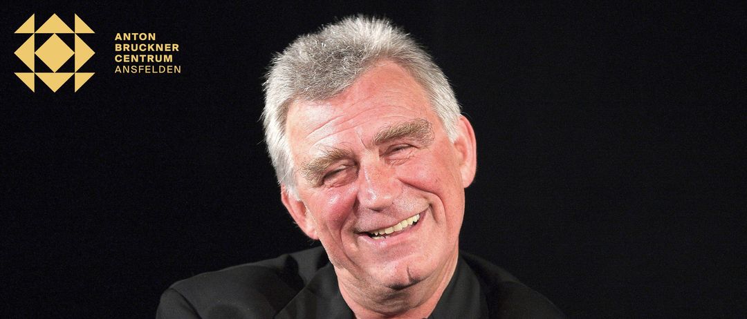 Ein Mann mit grauem Haar und einem Schnurrbart, der lächelt, ein schwarzes Hemd trägt und in ein Mikrofon spricht.