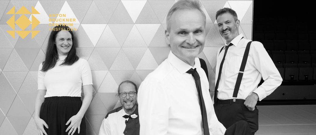 Ein monochromes Bild zeigt vier Erwachsene, die eng beieinander stehen. Drei Männer und eine Frau tragen formelle Kleidung. Die Männer lächeln und tragen Krawatten. Die Frau schaut nach rechts.