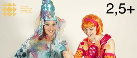 Zwei Frauen in bunten Kostümen, eine trägt einen blauen Hut, lächeln und halten einen Kuchen. Die Frau in Blau hält einen Kuchen auf einem silbernen Teller.