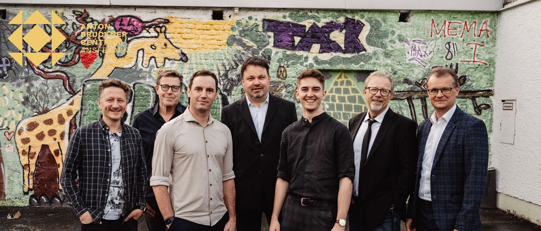 Eine Gruppe von Männern steht vor einer Graffiti-Wand, alle lächeln und posieren für ein Foto. Der Mann in der Mitte trägt ein schwarzes Shirt, während andere unterschiedliche Kleidungsstile tragen.