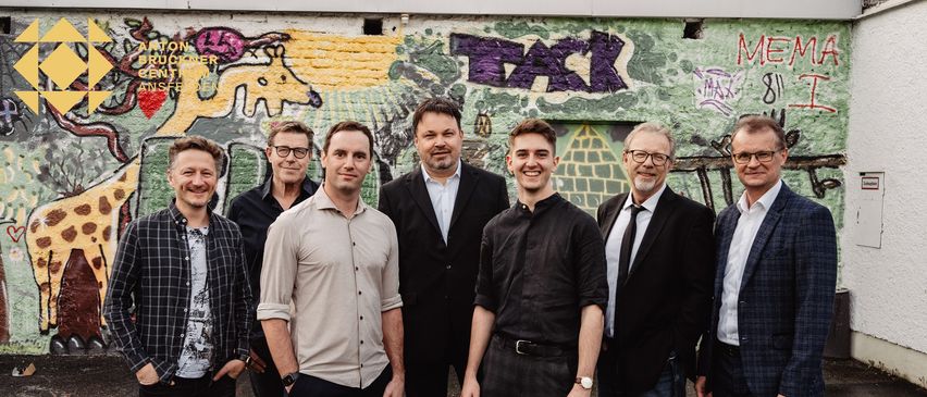 Eine Gruppe von Männern steht vor einer Graffiti-Wand, alle lächeln und posieren für ein Foto. Der Mann in der Mitte trägt ein schwarzes Shirt, während andere unterschiedliche Kleidungsstile tragen.