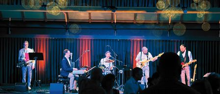 Eine Band spielt auf der Bühne mit blauen Vorhängen im Hintergrund. Der Leadsänger spielt Klavier, während ein anderes Mitglied Schlagzeug spielt. Der Gitarrist steht vor einem Mikrofon. Das Publikum ist unscharf.