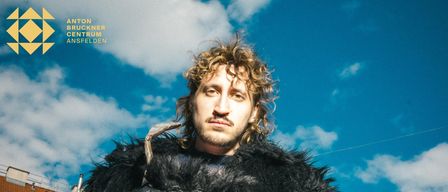 Ein Mann mit lockigem Haar, der einen schwarzen Pelzmantel und Lederhandschuhe trägt, steht vor einem blauen Himmel. Er hält ein Horn und schaut nach vorne.