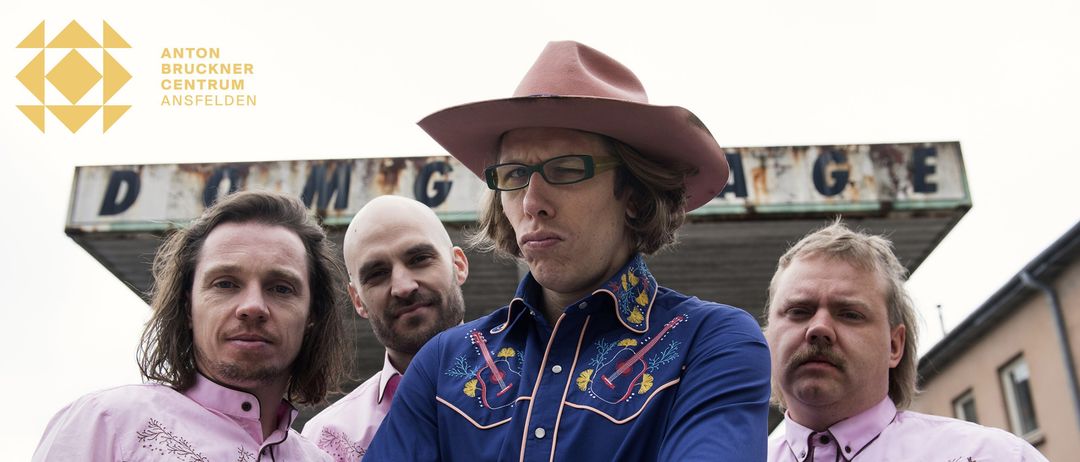 Vier Männer in Cowboy-Kleidung stehen draußen, einer mit Cowboyhut und Brille, einer kahlköpfig mit Schnurrbart und zwei in rosa Hemden.