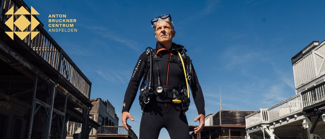 Ein älterer Mann in einem Taucheranzug steht vor einer Kulisse, die einer Westernstadt ähnelt. Der Himmel ist klar mit einigen Wolken.