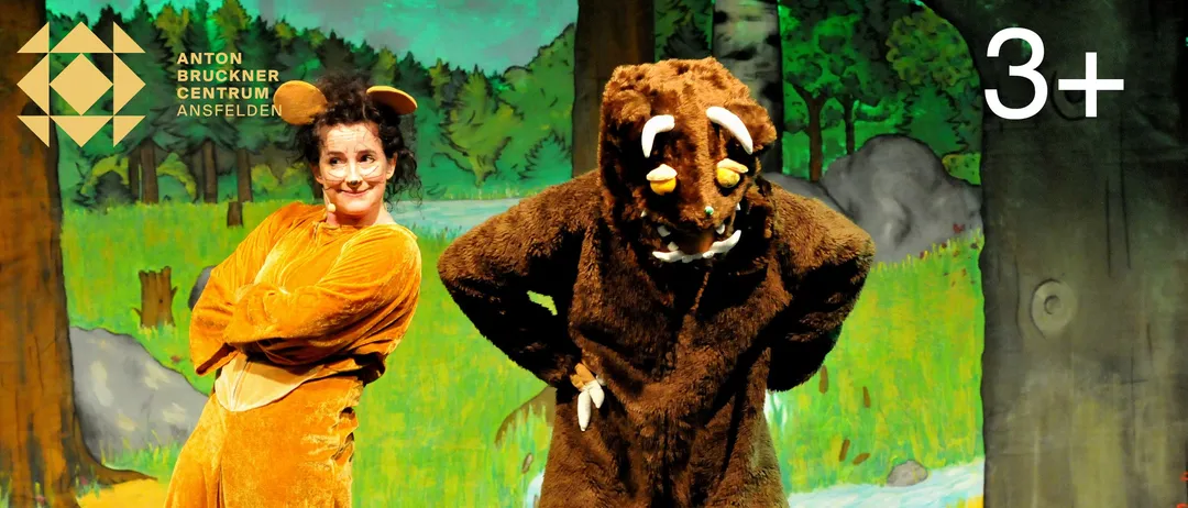 Zwei Darsteller stehen auf der Bühne, einer in einem Wolfskostüm und der andere in einem gelben Kostüm mit weißer Gesichtsmaske. Der Wolf steht mit den Händen auf den Hüften, und die Frau lächelt und posiert. Ein verschwommenes Bild von einer Kinderhand ist vor ihnen.