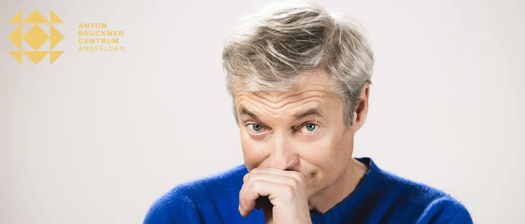 Ein Mann mit grauem Haar trägt einen blauen Pullover, schaut nach oben und hat die Hand vor dem Mund. Er scheint sich in einem Studio zu befinden.