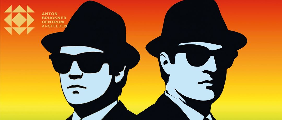 Zwei Männer in schwarzen Anzügen und Krawatten, beide tragen Hüte und Sonnenbrillen, stehen Seite an Seite vor einem farbenfrohen Hintergrund.