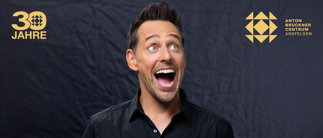 Ein Mann mit schwarzem Hemd und Bart lächelt und scheint zu lachen. Er hat blaue Augen und sieht nach oben.