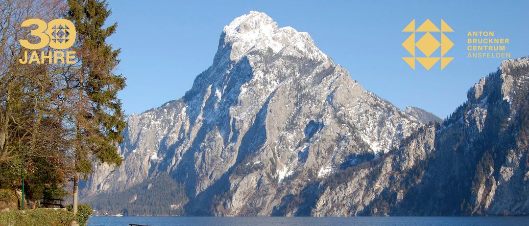 Ein Berg mit Schnee auf seinem Gipfel und felsiger Oberfläche mit einem Gewässer darunter unter einem klaren Himmel.